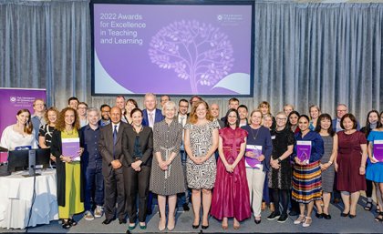 The winners of the 2022 UQ Awards for Excellence in Teaching and Learning. a group of people smiling on a stage in front of a big purple sign
